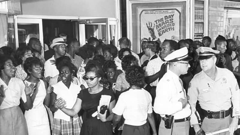 Picketing at the State Theater in Tallahassee in 1963; Patricia Due Stephens (in the black dress) and John Due (his head is visible behind the officer) are both pictured.