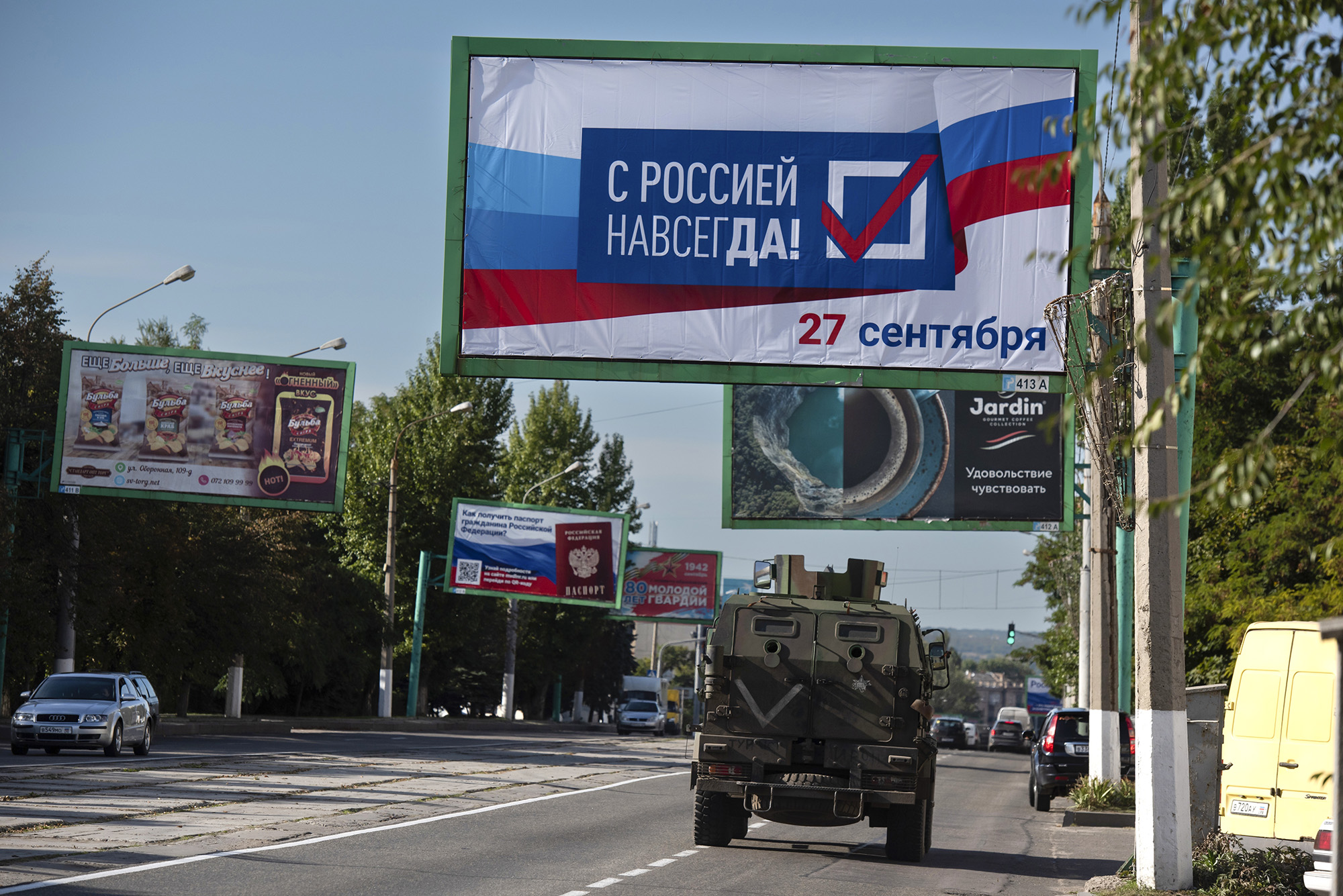 A military vehicle drives along a street with a billboard reading 