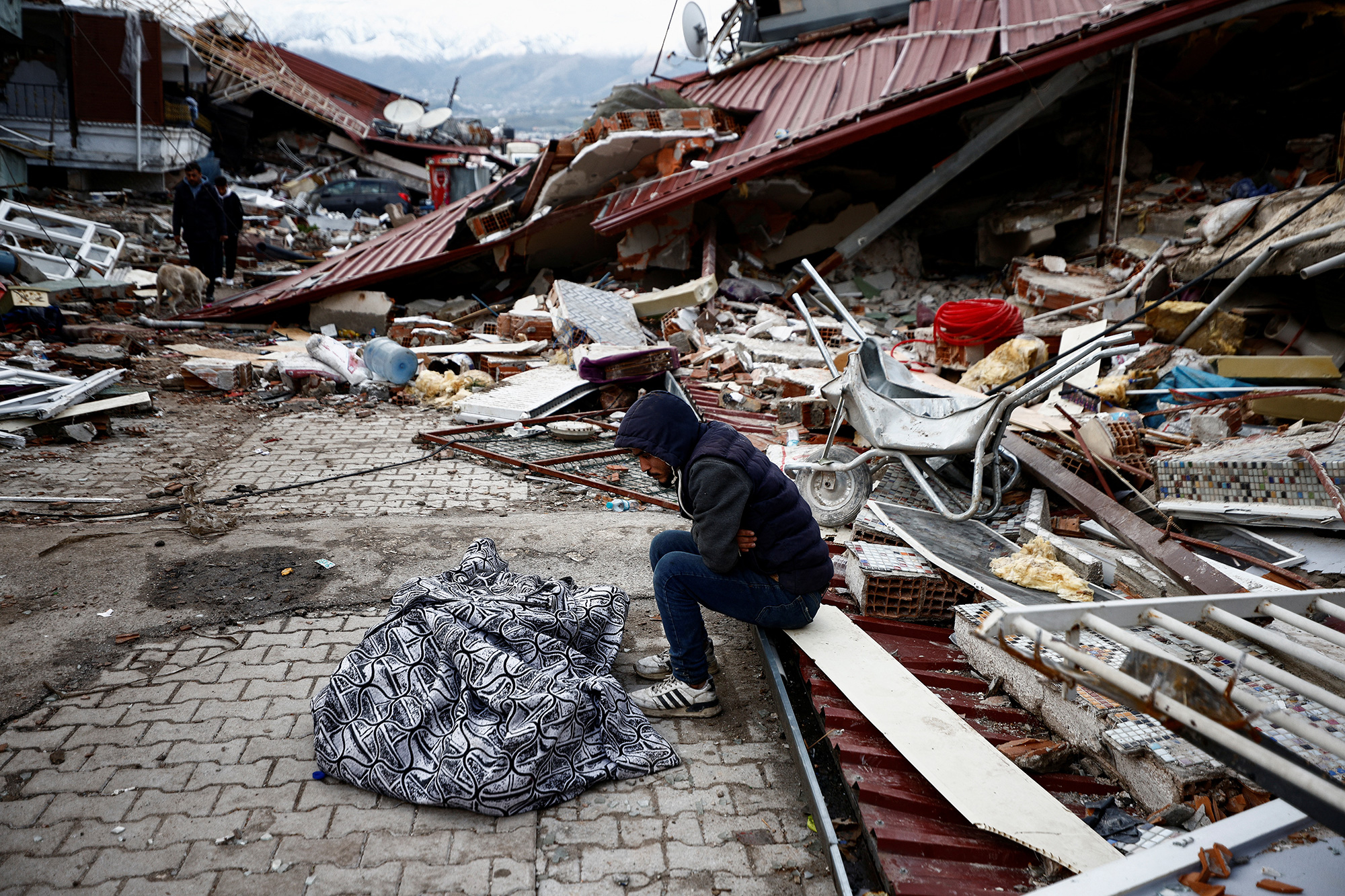 A man sits next to the body of a loved one covered with a blanket at the site of a collapsed building in Hatay, Turkey.