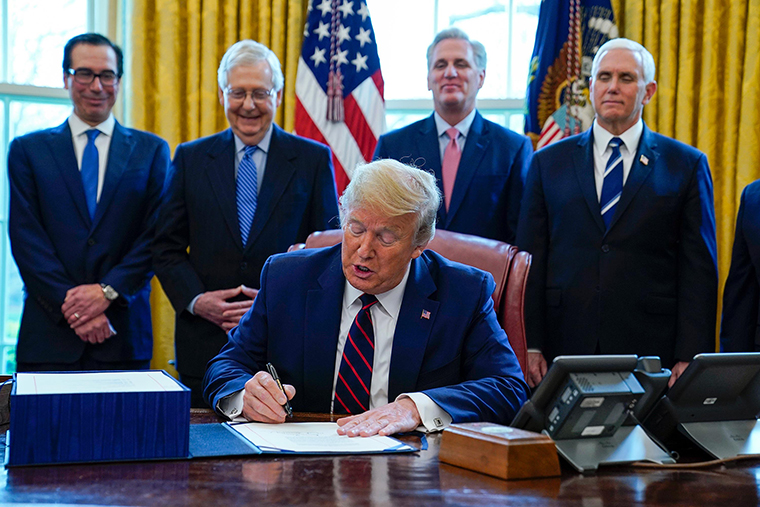 President Donald Trump signs the coronavirus stimulus relief package in the Oval Office at the White House, Friday, March 27, as Treasury Secretary Steven Mnuchin, Senate Majority Leader Mitch McConnell, House Minority Leader Kevin McCarty, and Vice President Mike Pence watch. 