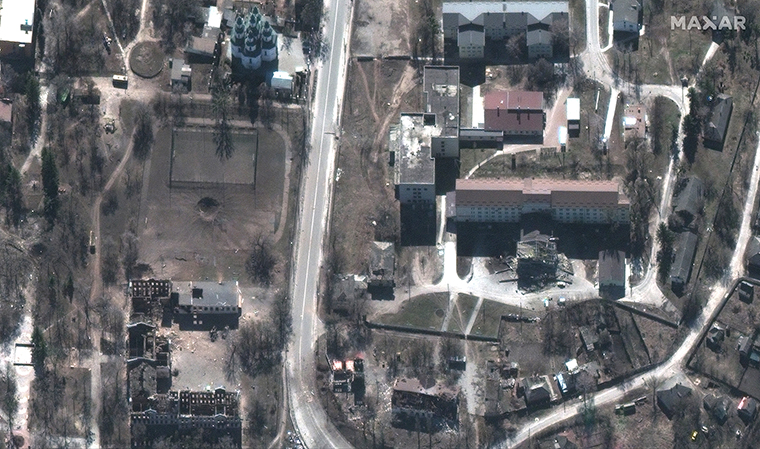 A satellite photo shows damage to izyum, Ukraine from Russian strikes.  A massive crater can be seen in the image, on one side of the crater is a school, and the other a soccer field. A damaged hospital can also be seen. 