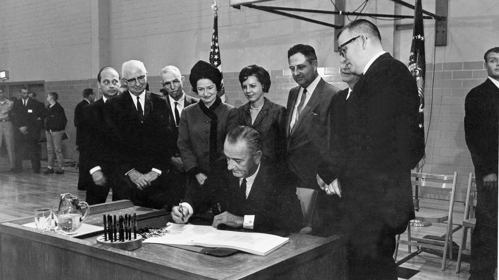 President Lyndon B. Johnson, seated at the desk he used while a student secretary at Southwest Texas State College 35 years ago, signs the Higher Education Act at San Marcos, Texas, in November 1965.