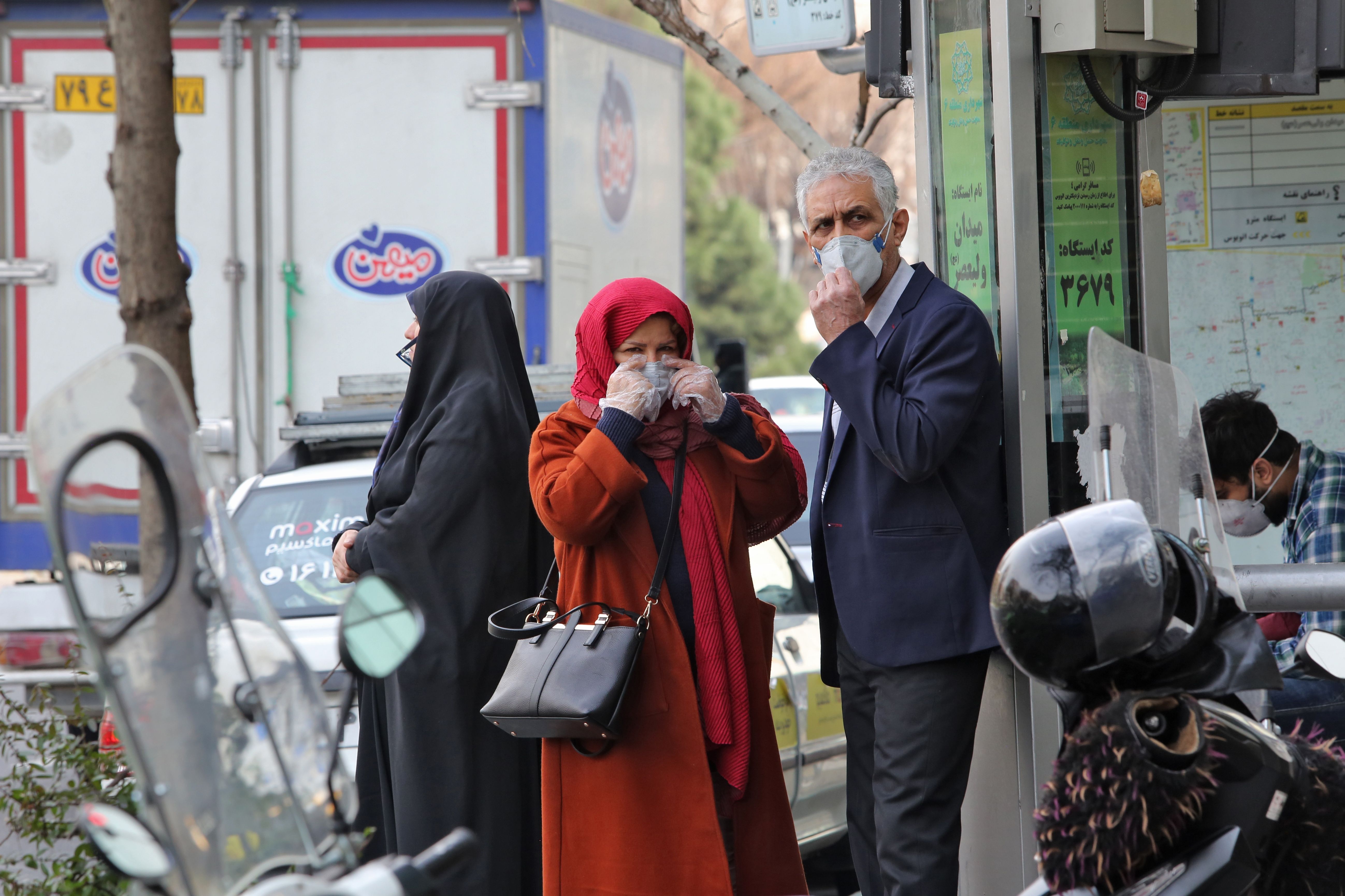 People wearing protective masks wait along the side of a street in the Iranian capital Tehran.