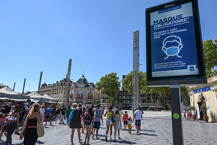 People wearing face masks walk next to a billboard reading "Mandatory mask in downtown Montpellier", on August 14, 2020, in Montpellier, southern France.