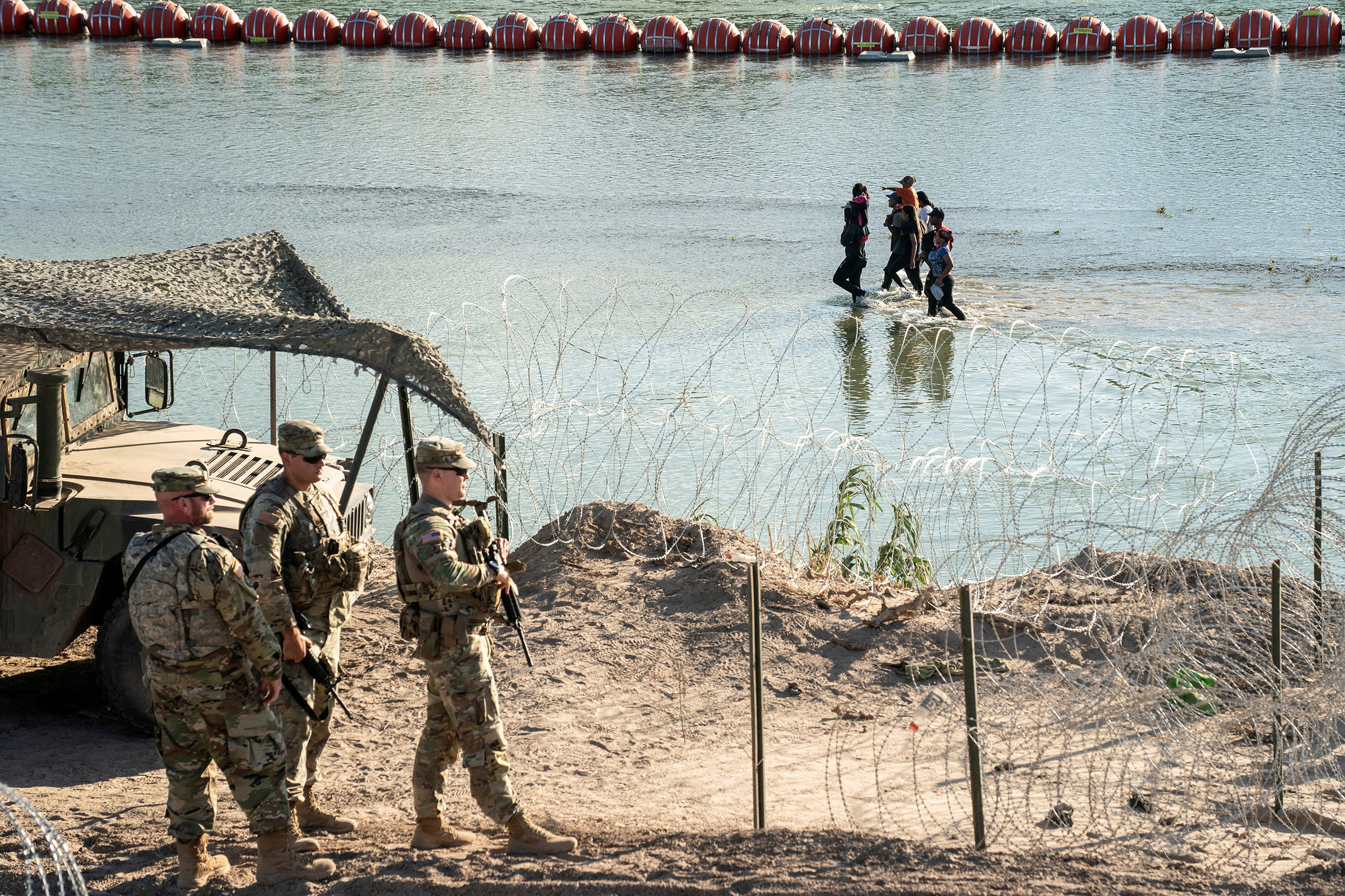 Asylum-seeking migrants walk in the Rio Grande river between a floating fence and the river bank as they look for an opening in a concertina wire fence to land on US soil in Eagle Pass, Texas, on July 24, 2023.
