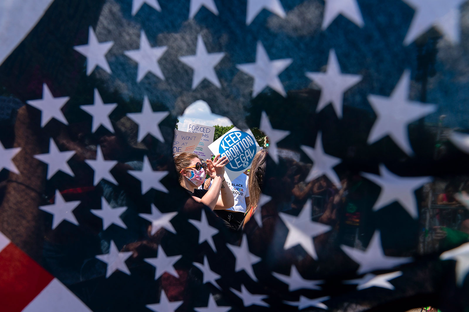 Abortion rights activists are seen through a hole in an American flag as they protest outside the Supreme Court in Washington, DC on Saturday, June 25.