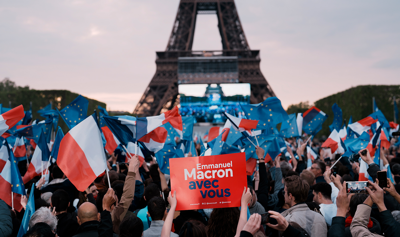 A supporter of French President Emmanuel Macron holds a placard reading "Emmanuel Macron With You" and others wave flags as they watch the first election projections being announced in Paris, France, Sunday, April 24.
