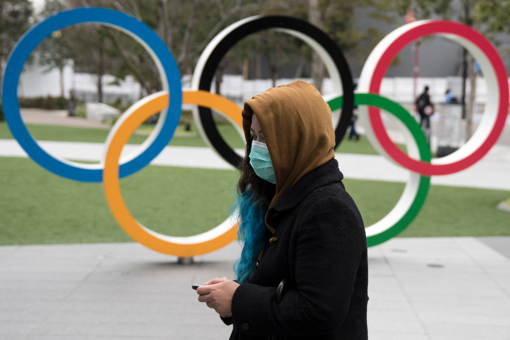 A woman wearing a face mask walks past the Olympic rings in front of the new National Stadium, the main stadium for the upcoming Tokyo 2020 Olympic and Paralympic Games, on February 26, 2020 in Tokyo, Japan. 