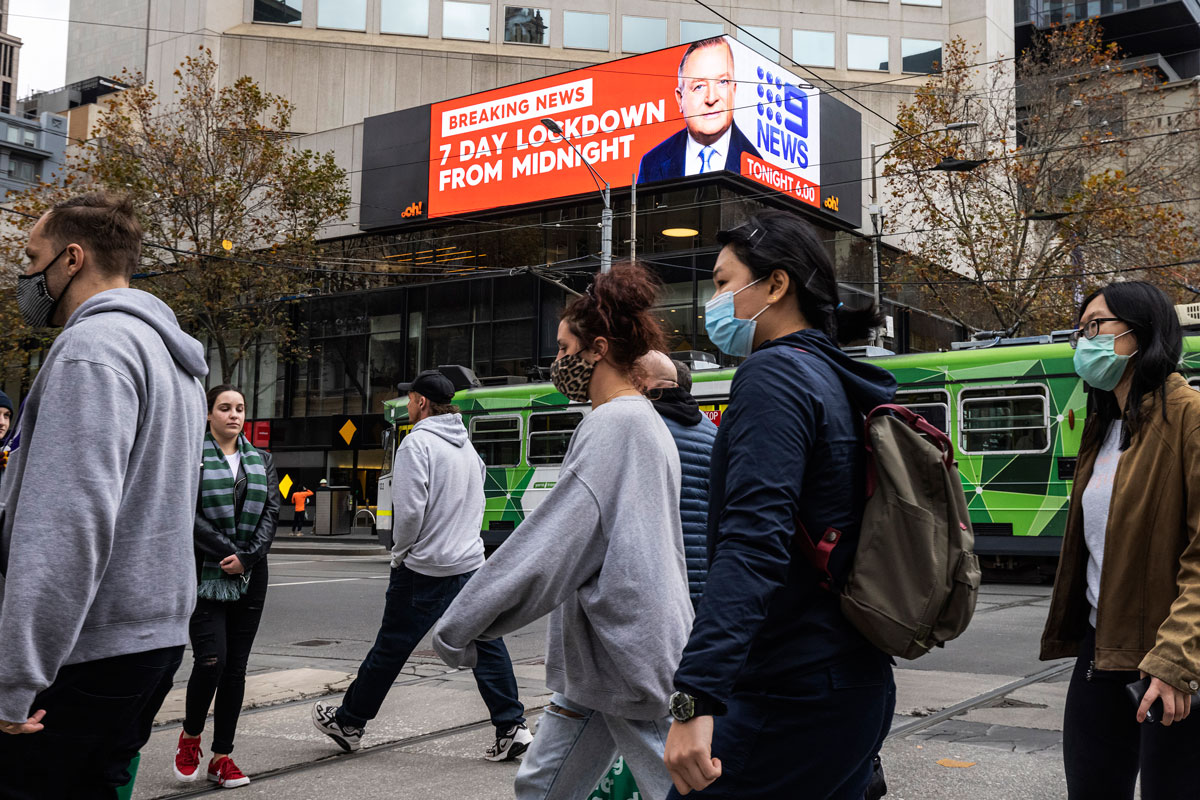 People wearing face masks are seen crossing Bourke Street Mall in front of an LED sign in the background displaying news of the impending seven-day lockdown on May 27 in Melbourne, Australia.