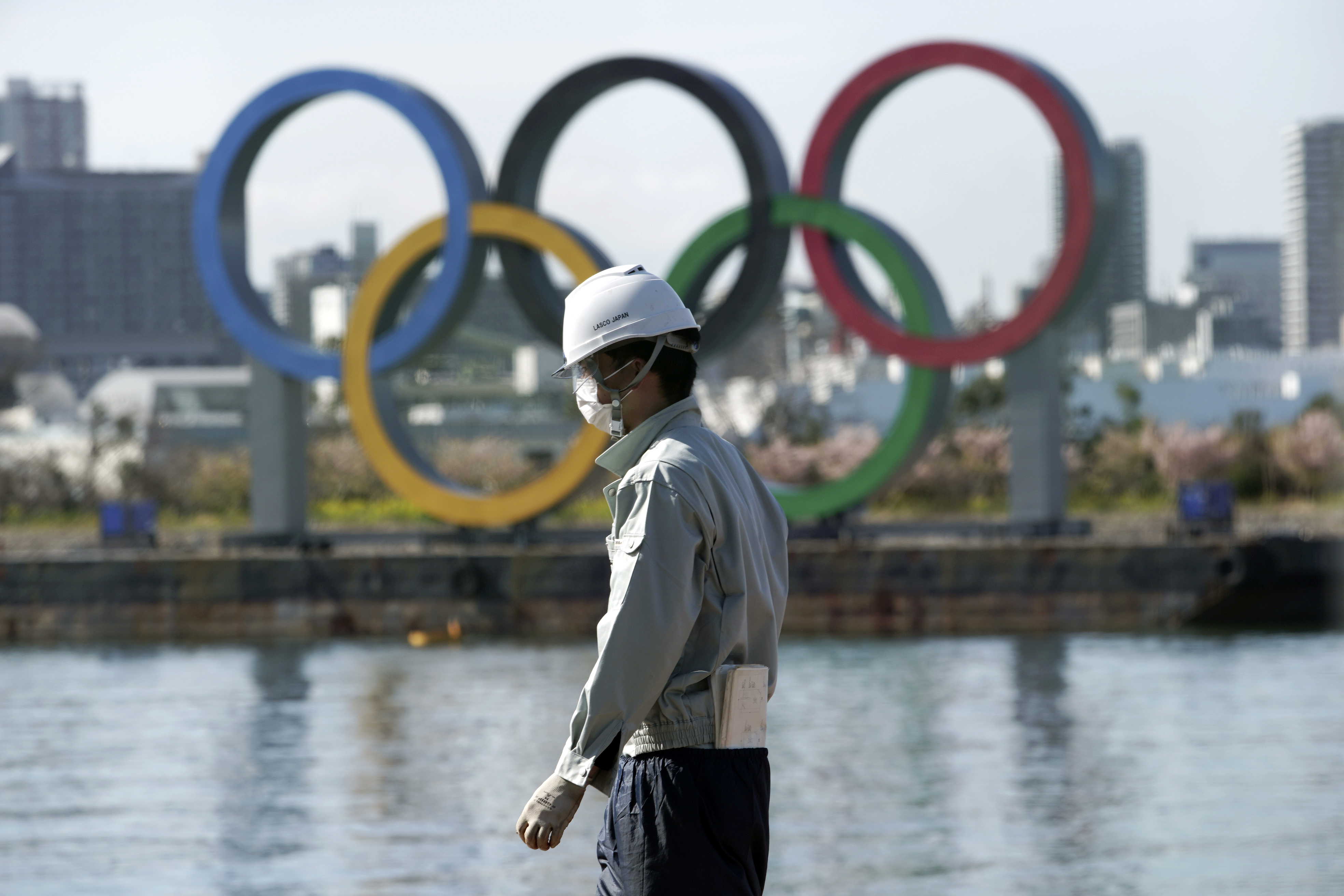 A masked man works at a construction site with the Olympic rings in the background Tuesday, March 3, in Tokyo.