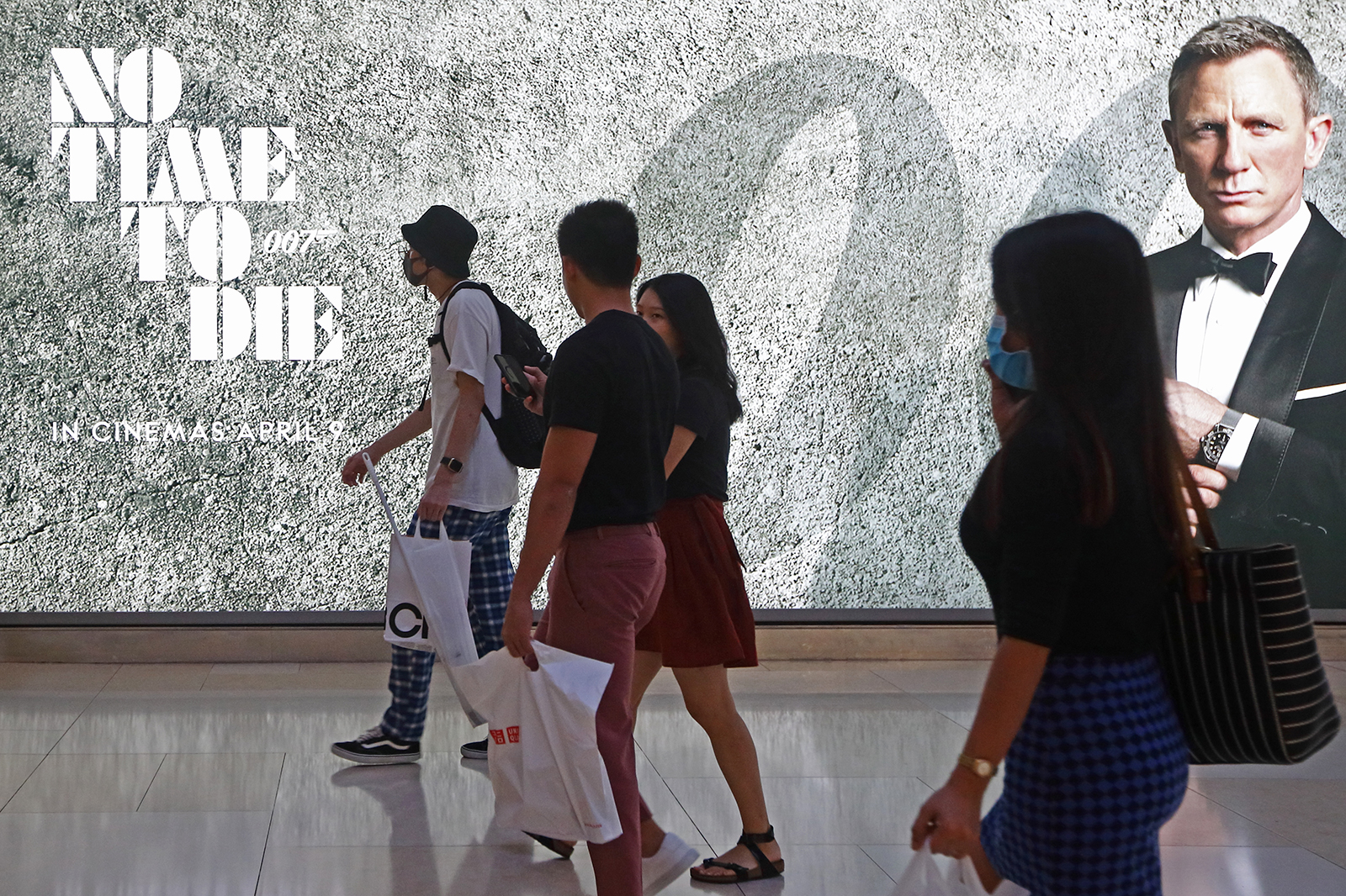 Shoppers wearing protective masks walk past a James Bond "No Time To Die" billboard in Singapore, on March 29, 2020.