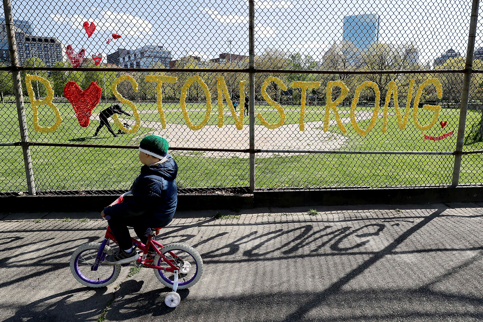 A child rides a bicycle with training wheels past a fence with the slogan "Boston Strong" crocheted into it, on Wednesday, May 13, in Boston.