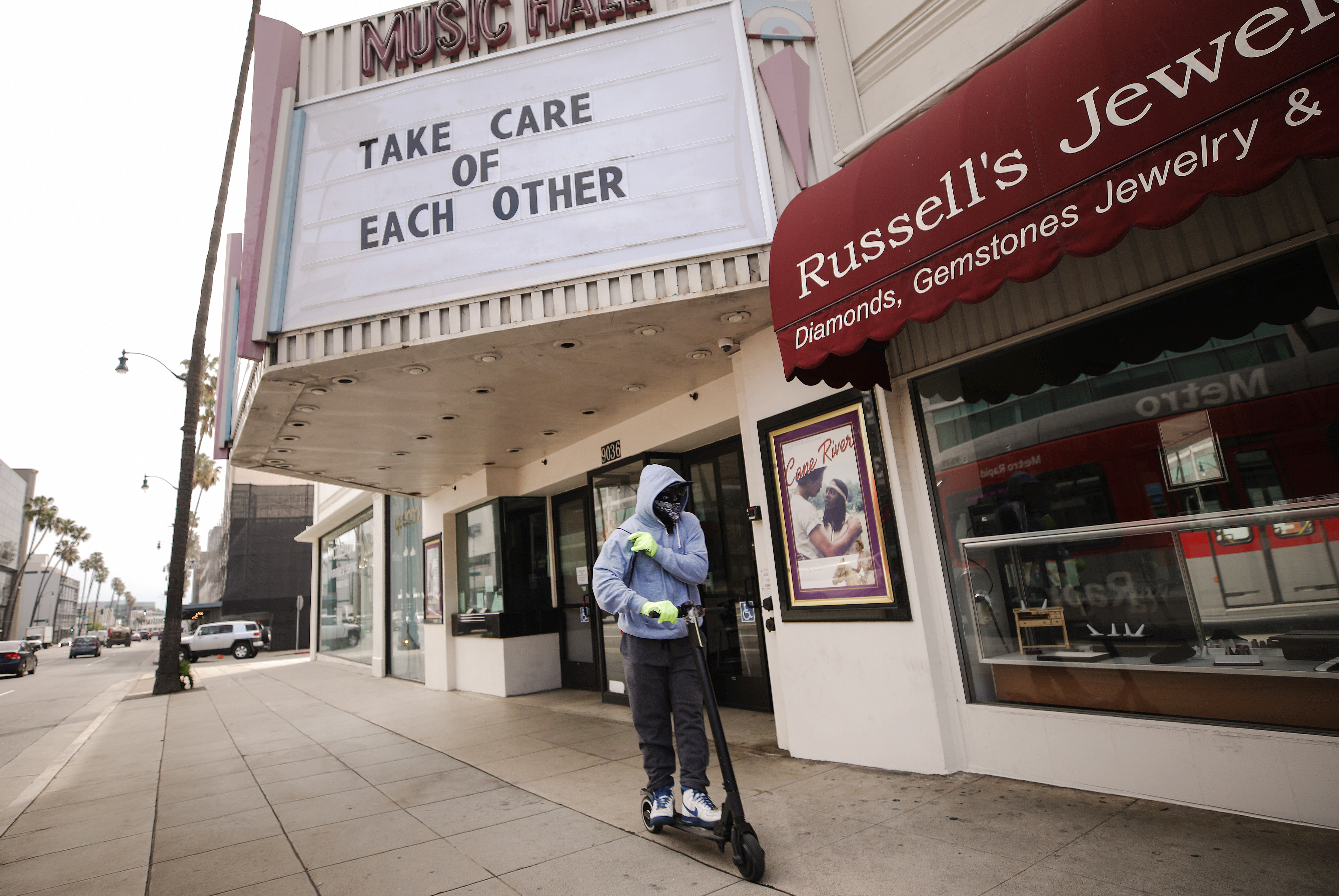 A man wears gloves and a bandanna across his face while riding a scooter past a shuttered movie theater, with the message "Take Care of Each Other" displayed on the marquee, on March 18, in Beverly Hills, California.