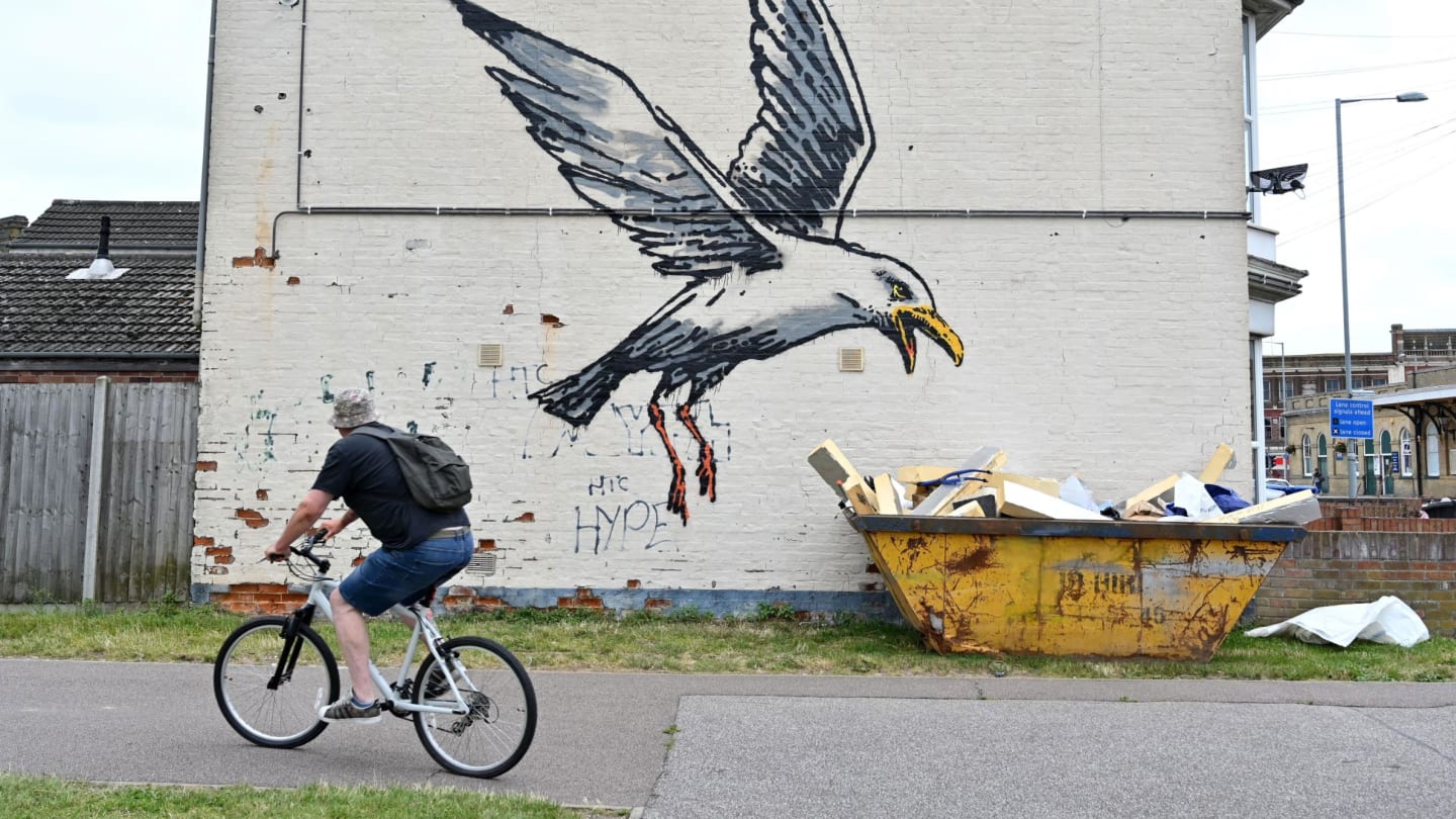 A cyclist rides past a stensil of a gull about to swoop down onto a carton of chips, the subject of a graffiti artwork bearing the hallmarks of street artist Banksy on a wall in Lowestoft on the East Coast of England on August 8, 2021. 